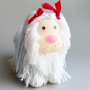 Puede incluir: Un juguete de peluche con forma de perro con una cara clara, una nariz rosa y ojos negros. El juguete tiene pelo de hilo blanco y azul y una cinta roja. El perro está sobre una superficie gris.