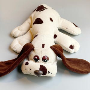 Könnte beinhalten: Ein Vintage-Plüschtier in Form eines Hundes, cremefarben mit braunen Flecken und Schlappohren. Der Hund liegt ausgestreckt, mit großen, ausdrucksstarken Augen und einer braunen Nase. Ein weiches und kuscheliges Spielzeug für Kinder oder Sammler.