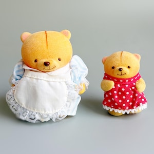 May include: Two yellow bear figurines. One bear wears a blue and white dress with a white apron. The other bear wears a red dress with white polka dots. Both bears have small, black eyes and brown noses.
