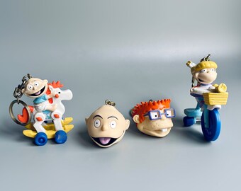 May include: Four plastic keychains featuring characters from the Nickelodeon cartoon Rugrats. The keychains feature Tommy Pickles riding a rocking horse, Chuckie Finster's head, Phil and Lil DeVille's head, and Angelica Pickles riding a tricycle.