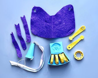 Könnte beinhalten: Ein lila Filzumhang, vier lila Filzstreifen, eine blaue Plastik-Partyhupe, eine blaue Plastikkrone mit goldenen Akzenten, zwei goldene Plastikstäbe und ein goldener Plastikring. Alle diese Gegenstände liegen auf einem hellblauen Hintergrund.