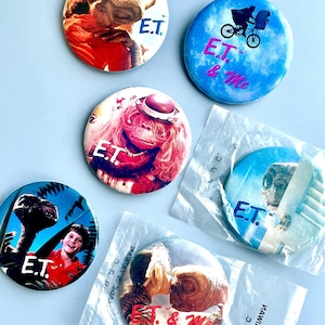 Puede incluir: Un conjunto de botones redondos con imágenes y texto de la película "E.T. el Extraterrestre". Los botones muestran varias escenas y personajes, incluyendo a E.T. y los textos "E.T." y "E.T. & Me."