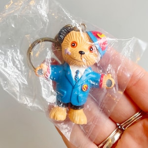 May include: A small, vintage keychain featuring a bear character in a blue suit with a red and blue hat. The bear has a gold keychain ring attached and is inside a clear plastic bag. The bear's suit has a yellow "S" on the chest.