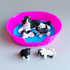 Pode incluir: Uma coleção de estatuetas de gatos. Uma estatueta de gata mãe com marcas em preto e branco repousa em uma cama de plástico rosa com três gatinhos. Duas estatuetas de gato adicionais também estão presentes, uma em preto e branco e outra em preto sólido.