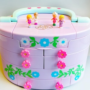 May include: A vintage Polly Pocket playset in a lilac case with a handle. The case is decorated with floral designs and pink flower-shaped drawer pulls. Four miniature figures stand on top.