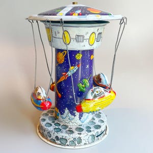 Puede incluir: Un tiovivo de juguete de hojalata vintage con temática espacial. El tiovivo presenta una torre azul y blanca con un diseño de cielo estrellado, y tres asientos con forma de nave espacial colgando de alambres. El tiovivo está pintado con cohetes, planetas y estrellas.