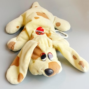 May include: A plush dog toy in cream with brown spots. The dog has long floppy ears, black and red eyes, and a black nose. A red and white button is on the dog's back. The toy is lying on a gray surface.