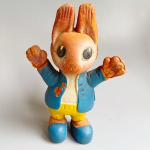 Puede incluir: Una figura de conejo de cerámica vintage con los brazos extendidos. El conejo tiene orejas naranjas, una cara color melocotón y lleva una chaqueta azul sobre una camisa blanca y pantalones amarillos. Zapatos azules completan el atuendo.