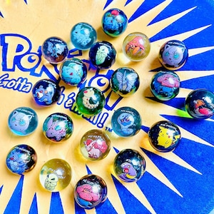 Peut inclure: Une collection de 18 billes en verre colorées représentant des personnages Pokémon. Les billes sont disposées en cercle sur un fond bleu et jaune avec le texte "Gotta catch 'em all!"