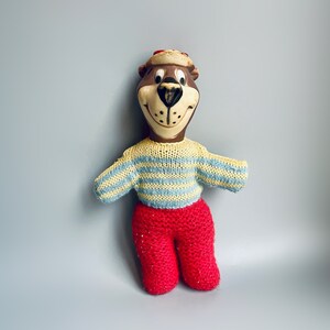 Puede incluir: Muñeca de oso vintage con cabeza marrón, suéter a rayas amarillas y azules y pantalones de punto rojos. El oso tiene una cara sonriente y un sombrero pequeño. La muñeca está sobre un fondo azul claro.