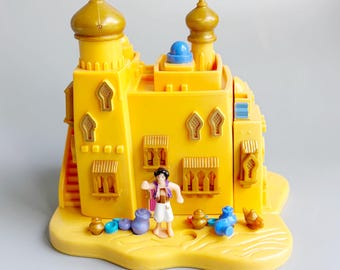 Può includere: Un castello giocattolo giallo con dettagli dorati, con una piccola figura e altri accessori in miniatura. Il castello ha più torri e finestre, posto su una base sabbiosa. È presente anche una figura di genio blu e altri piccoli oggetti.