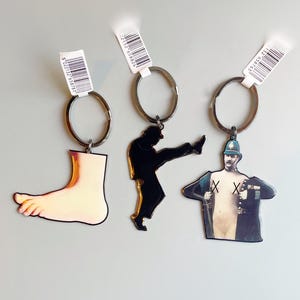 Może przedstawiać: Trzy breloczki do kluczy z srebrnymi kółkami. Jeden breloczek przedstawia stopę, drugi czarną sylwetkę kopiącej osoby, a trzeci obrazek policjanta w stylu vintage. Każdy breloczek ma etykietę z kodem kreskowym.