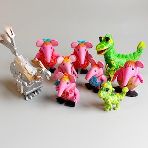 Puede incluir: Colección de figuras coloridas, que incluyen criaturas rosas parecidas a ratones con lazos azules y ropa estampada, un dinosaurio verde, un pequeño dinosaurio amarillo y verde y una figura mecánica plateada. Los juguetes están dispuestos sobre una superficie blanca.