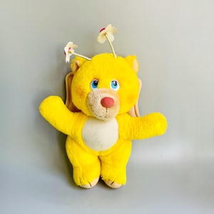 Op de afbeelding: Een gele pluche teddybeer met vleugels en bloemen op zijn kop. De beer heeft blauwe ogen en een roze neus.