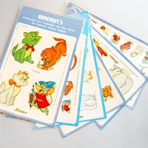 May include: A stack of vintage Bancroft's art transfer sheets featuring colorful cartoon cat and mouse characters. The sheets are in plastic packaging with blue borders and text that reads "Colourful Art Transfers for the Home by famous Italian Artists."