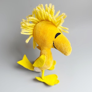 Puede incluir: Un personaje de p&aacute;jaro de peluche amarillo con un pico grande y ojos negros. El p&aacute;jaro tiene un mech&oacute;n de hilo amarillo en la cabeza y est&aacute; de pie sobre dos pies amarillos. El personaje es de la tira c&oacute;mica Peanuts.