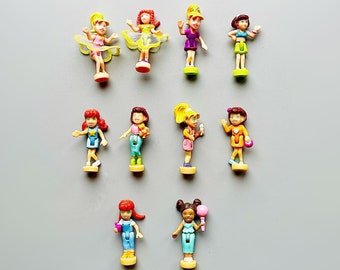 Könnte beinhalten: Eine Sammlung von zwölf kleinen Plastikpuppen, jede mit einzigartigen Outfits und Frisuren. Die Puppen befinden sich in verschiedenen Posen, einige halten Zubehör. Die Farbpalette umfasst Rosa, Gelb, Blau und Orange, vor einem grauen Hintergrund.