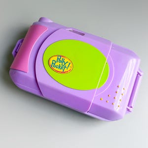 Könnte beinhalten: Ein Vintage Polly Pocket Kompaktspielzeug in rechteckiger Form. Das Spielzeug ist hauptsächlich lila mit rosa und grünen Akzenten. Das Polly Pocket-Logo ist auf der Vorderseite zu sehen. Kleine gelbe Punkte an der Seite.