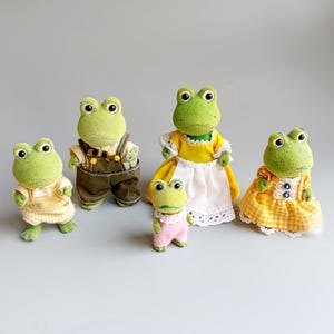 以下が含まれることがあります： さまざまな衣装を着た5匹のカエルの置物。カエルは、緑、黄色、茶色、ピンクなど、さまざまな色があります。衣装には、オーバーオールやドレスが含まれます。置物は、明るい灰色の表面に配置されています。