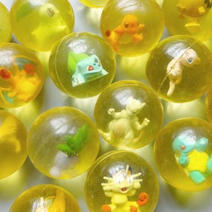 Peut inclure: Collection de jouets sphériques transparents jaunes, chacun contenant une petite figurine de personnage colorée. Les figurines sont des personnages Pokémon variés. Les jouets sont disposés de près, remplissant le cadre.