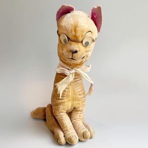 May include: Vintage plush dog toy with oversized googly eyes, pink-lined ears, and a cream-colored bow. The dog is a light tan color and has a stitched black nose. The toy is sitting upright, with its legs extended.