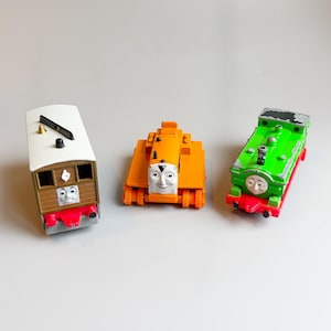 May include: Three toy train engines: a brown and white train car, an orange engine, and a green engine. Each train has a smiling face on the front. The brown train has a white roof and a black bell.