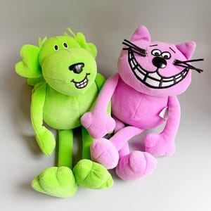 以下が含まれることがあります： 2つのぬいぐるみ：緑色のライオンとピンクの猫。ライオンは笑顔で、緑色のたてがみと緑色の足を持っています。猫は大きな笑顔、黒いひげ、ピンクの足を持っています。どちらのおもちゃも柔らかく、抱きしめたくなります。