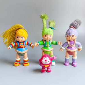 May include: Three plastic dolls with yarn hair and a small pink creature figure. The left doll has yellow hair and a blue outfit. The centre doll has green hair and a green outfit. The right doll has purple hair, glasses, and a purple outfit.
