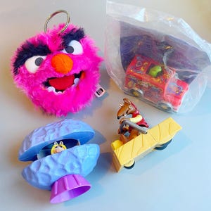 May include: A selection of Muppet Show character toys. Features a pink and black fuzzy Animal keyring, a red truck toy, a blue clam shell with a Miss Piggy figurine, and a yellow cart with a Gonzo figurine.