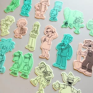 Puede incluir: Una colección de recortes de personajes de dibujos animados en varios colores, incluyendo verde, melocotón y azul. Los personajes están delineados en negro y parecen ser de una popular serie de televisión animada. Los recortes están dispuestos sobre una superficie gris claro.