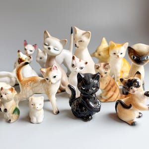 May include: A selection of ceramic cat ornaments in diverse poses and colours. The cats are white, orange, and black, some with detailed markings. The figurines vary in size and style, displaying a vintage aesthetic.