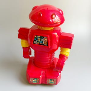 May include: A bright red plastic robot toy featuring a rounded head with two orange circular eyes, yellow accordion-style neck and arm joints, and a rectangular chest panel with a colorful graphic display. The base also features two small rectangular graphic panels.