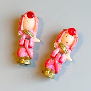Op de afbeelding: Twee kleine, vintage beeldjes van een meisje in een roze jurk met een rode edelsteen op haar hoofd. De beeldjes hebben gouden accenten op hun schoenen en armen. De achtergrond is effen lichtgrijs.
