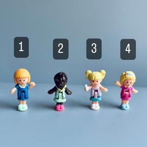 Może przedstawiać: Cztery miniaturowe plastikowe lalki. Lalka 1 ma blond włosy i niebieską sukienkę. Lalka 2 ma ciemną skórę i zielony strój. Lalka 3 ma blond warkocze i fioletowy strój. Lalka 4 ma blond włosy i różową sukienkę. Każda lalka ma około 3 cm wysokości.