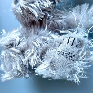 May include: A gray plush toy with a signature on the paw. The toy is made of soft, fluffy material.