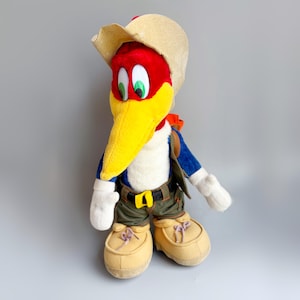 May include: A plush Woody Woodpecker doll wearing a tan cowboy hat, a blue jacket, and olive green shorts. The doll has a red head, a large yellow beak, and green eyes. It is wearing tan shoes and a brown belt with a gold buckle.