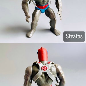 May include: A gray action figure of Stratos from the Masters of the Universe toy line. He is wearing a red helmet, blue goggles, a gray and white harness, and a blue and red loincloth. The figure is standing with its arms outstretched and its legs slightly apart. The figure is posed in a dynamic action pose. The figure is in good condition and has no missing parts.