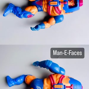 May include: A blue and orange action figure of Man-E-Faces from the Masters of the Universe toy line. The figure has a blue helmet with a face on it and a red and orange body. The figure is lying on its back with its arms and legs spread out.