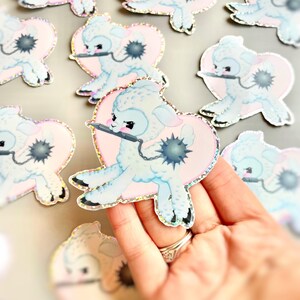 May include: A set of pastel blue and pink holographic stickers featuring a cartoon lamb holding a knife. The lamb is sitting in a heart shape with a black and white spiky ball in the background.