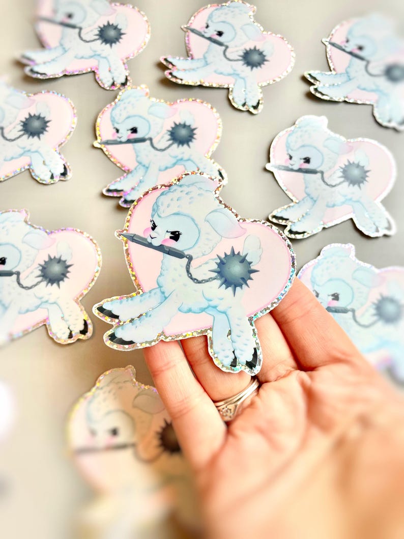 May include: A set of pastel blue and pink holographic stickers featuring a cartoon lamb holding a knife. The lamb is sitting in a heart shape with a black and white spiky ball in the background.
