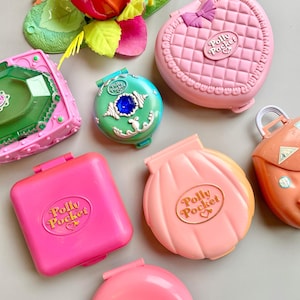 May include: A collection of seven plastic Polly Pocket toy cases in various shapes and colors, including a pink heart-shaped case, a shell-shaped case, a rose-shaped case, a bright pink square case, and a teal round case with a blue gem.