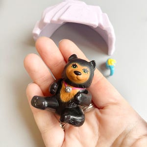 May include: A small black bear figurine with brown eyes and a pink collar with a silver charm. The bear is sitting up with its legs outstretched.