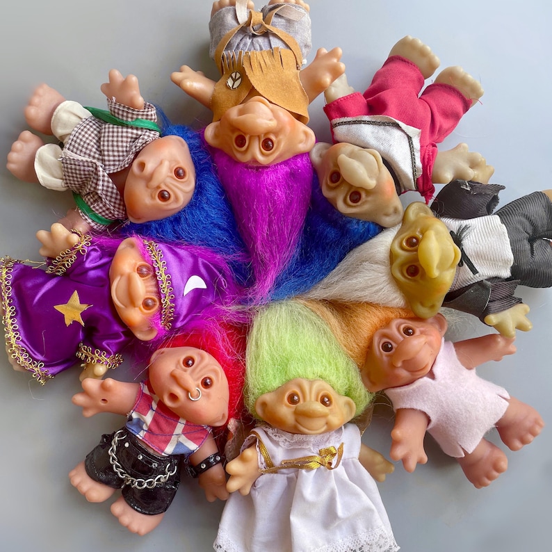 May include: A group of nine colorful troll dolls with different hairstyles and outfits. The dolls are arranged in a circle, with their heads facing the center. The dolls are made of plastic and have a variety of colors, including pink, blue, green, purple, and red.