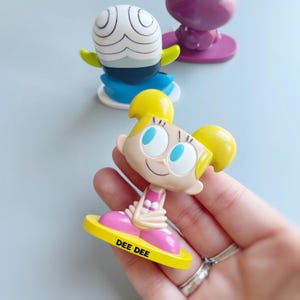 May include: A yellow and pink plastic figurine of Dee Dee from the Powerpuff Girls cartoon. Dee Dee is sitting on a yellow base with the words "DEE DEE" printed on it.