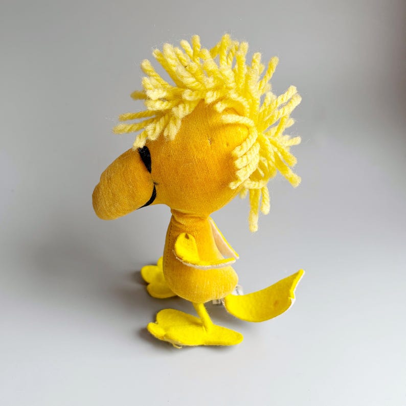 Puede incluir: Un peluche amarillo de Woodstock, un personaje de la tira c&oacute;mica Peanuts. El juguete tiene un pico amarillo grande, ojos negros y un mech&oacute;n de pelo de hilo amarillo. El p&aacute;jaro est&aacute; de pie sobre dos pies amarillos.