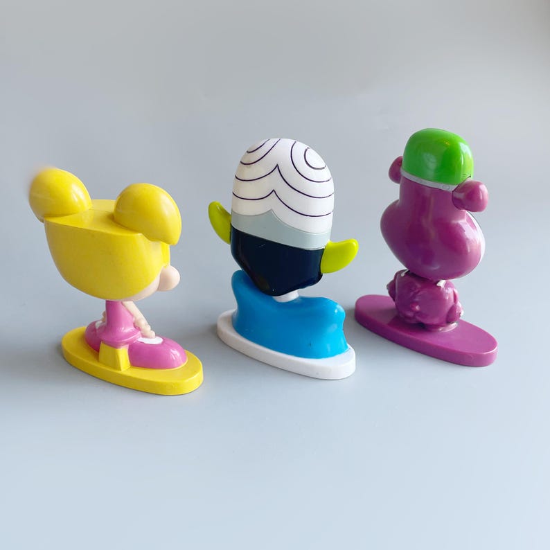 May include: Three cartoon character figurines, a yellow girl with pigtails, a black and white character with a green and blue base, and a purple character with a green hat. The figurines are on a white surface.