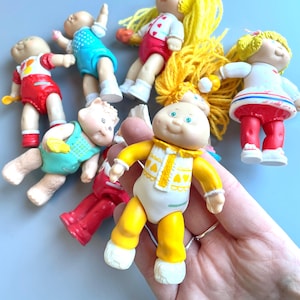 Peut inclure: Une collection de poupées Cabbage Patch Kids vintage, dont une poupée vêtue de jaune avec un motif en forme de cœur, une poupée vêtue de rouge avec une chemise à rayures blanches et bleues, une poupée vêtue de bleu avec une chemise blanche et une poupée vêtue de rose avec une chemise blanche.