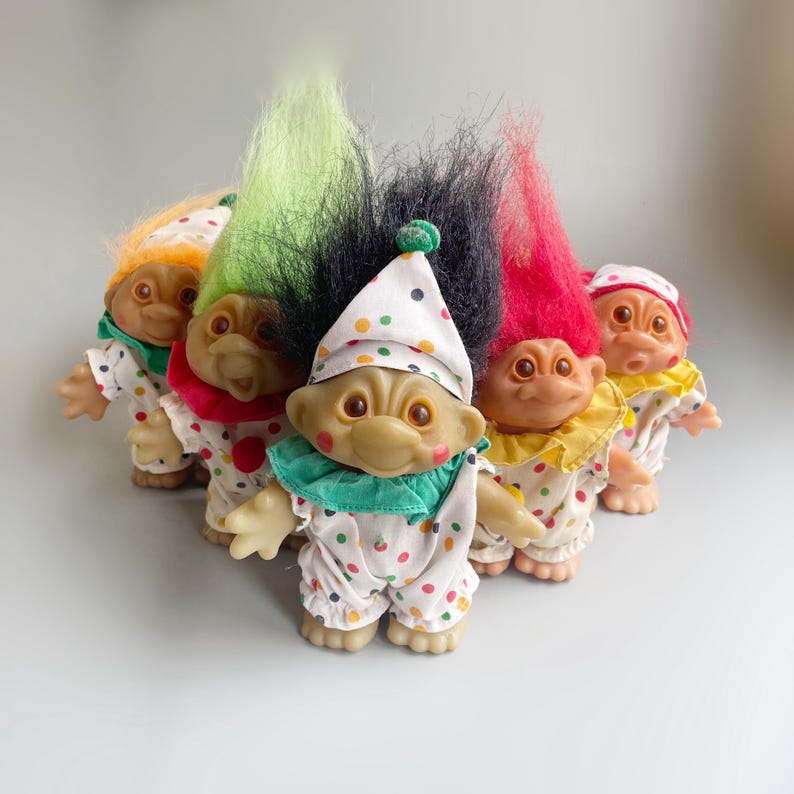 Pu&ograve; includere: Cinque bambole troll vestite da clown con abiti e cappelli a pois colorati. I troll hanno i capelli di diversi colori e sono disposti in fila.