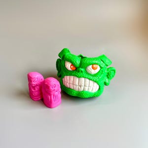 Könnte beinhalten: Ein grüner Plastikmonsterkopf mit einem zahnigen Grinsen und orangefarbenen Augen. Zwei rosa Plastikmonsterfiguren mit kleinen Armen und Beinen.