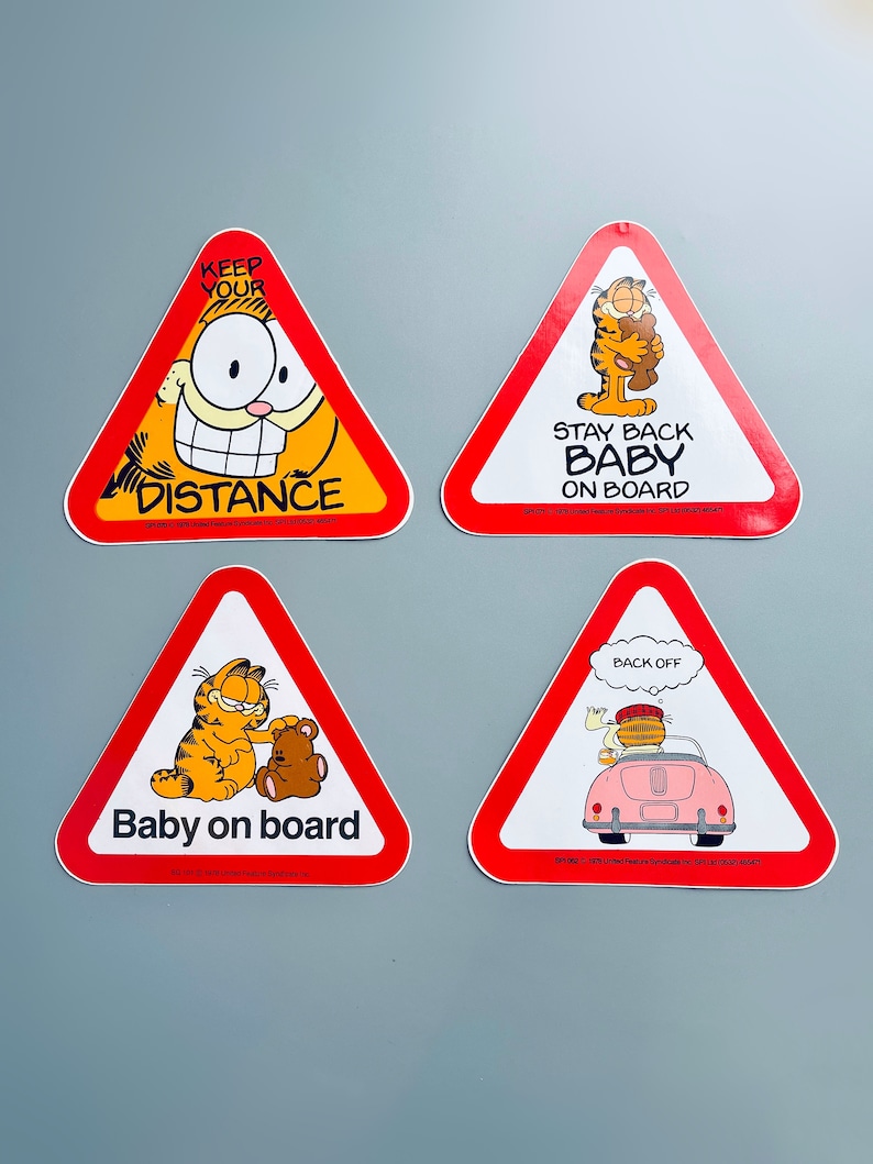 Puede incluir: Cuatro pegatinas de coche de forma triangular roja con un personaje de gato de dibujos animados, Garfield. Las pegatinas dicen "Keep Your Distance", "Stay Back Baby On Board", "Baby On Board", y "Back Off".